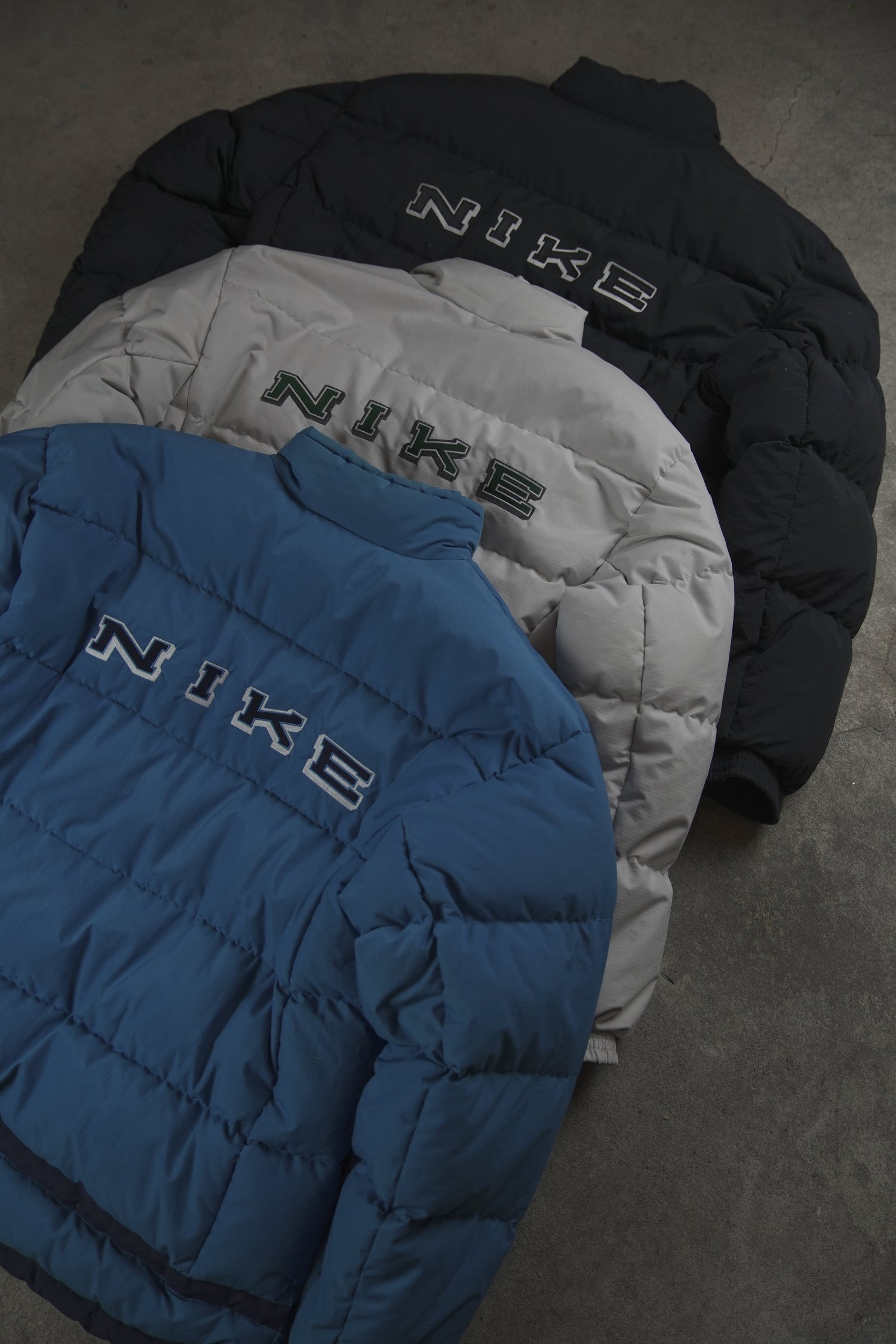 A stylized photo of several dark vintage Nike puffer jackets with a prominent logo, laid out on a gray surface, part of Atelier Solitaire's exclusive collection.